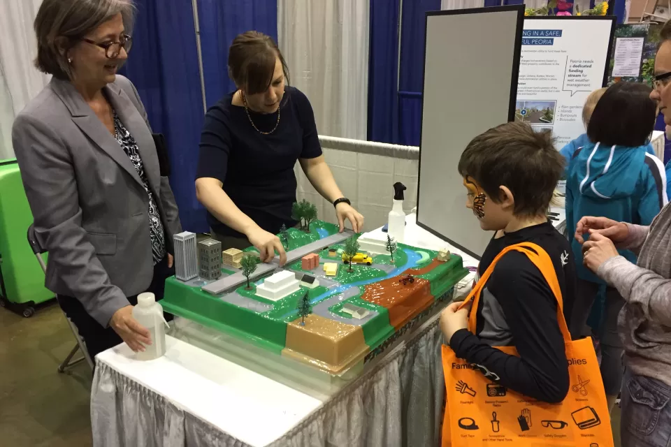Two adults stand behind a table, gesturing toward a model of a city while talking to a young child about buildings and parks and streets.